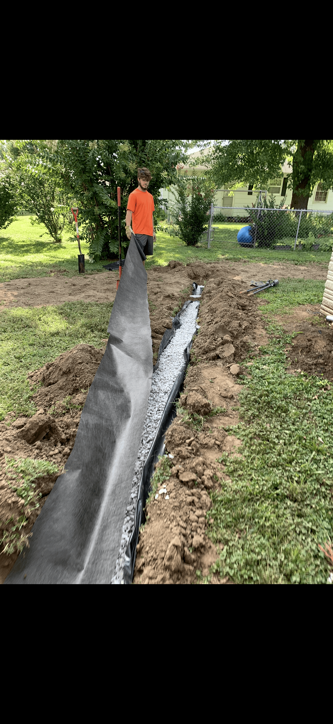 Man in orange shirt laying black landscape fabric over a gravel trench in a yard.