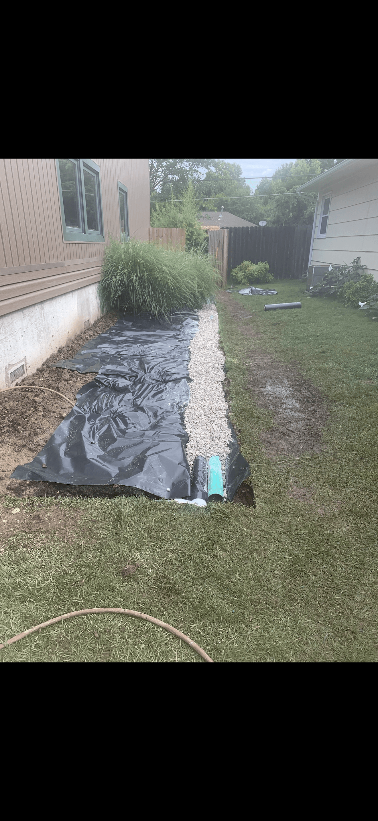 Backyard drainage installation featuring black plastic sheeting, white gravel, and a green drainage pipe.